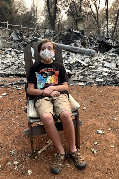 Child wearing a face mask sits on a chair amid charred debris from a recent fire.