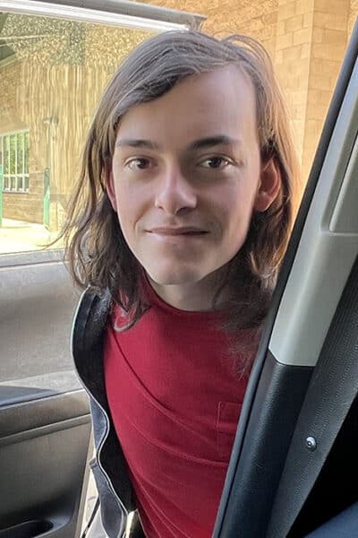 Young person with shoulder-length hair leaning into an open car door, wearing a red shirt, outdoors by a brick building.